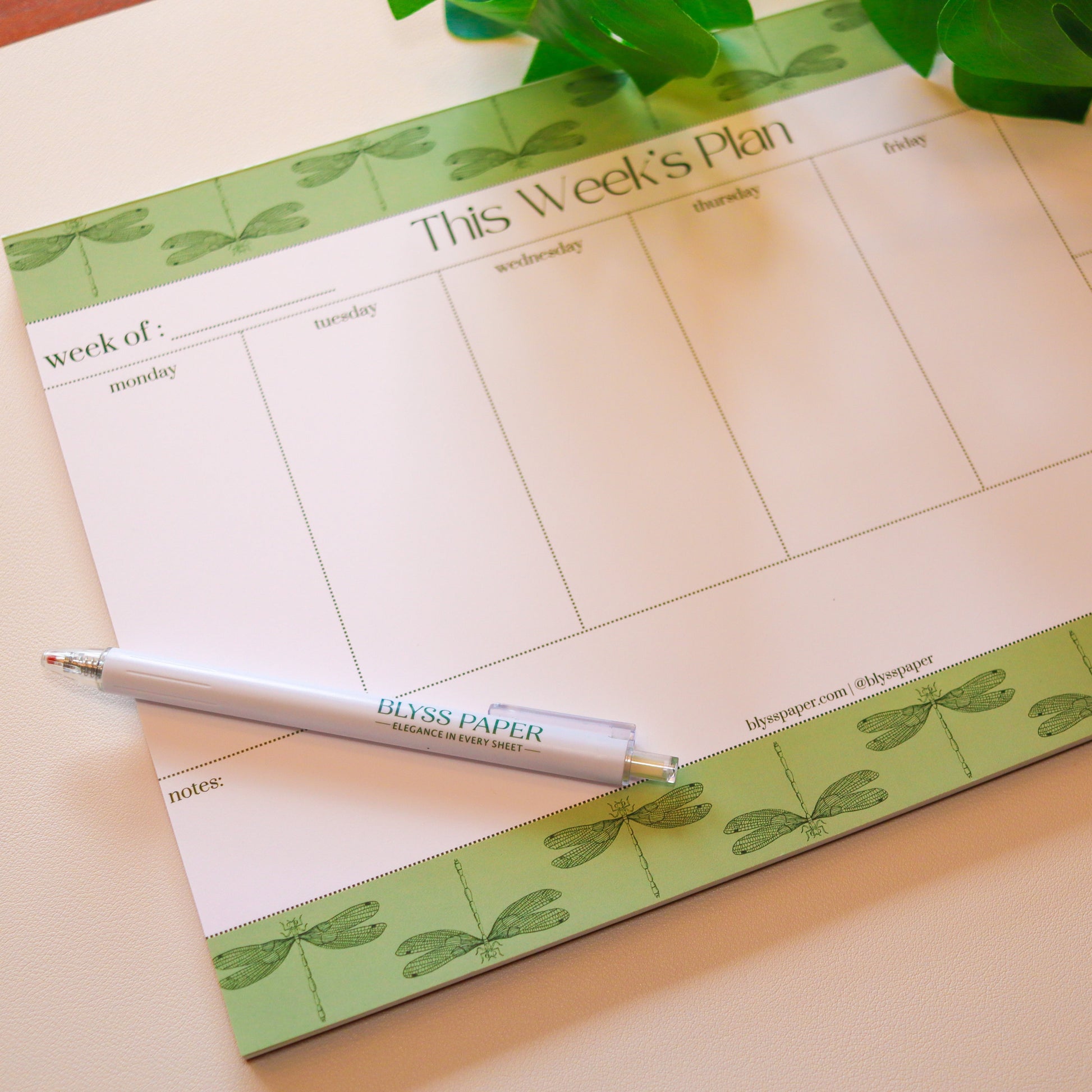 Weekly planner pad with a pen on a light surface, featuring green decorative elements.